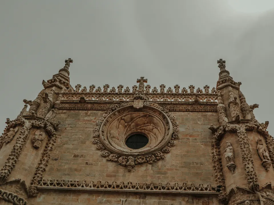 Carved Manueline stone facade with a round rose window and pinnacles at the Convent of Christ in Tomar, seen from below against a grey sky